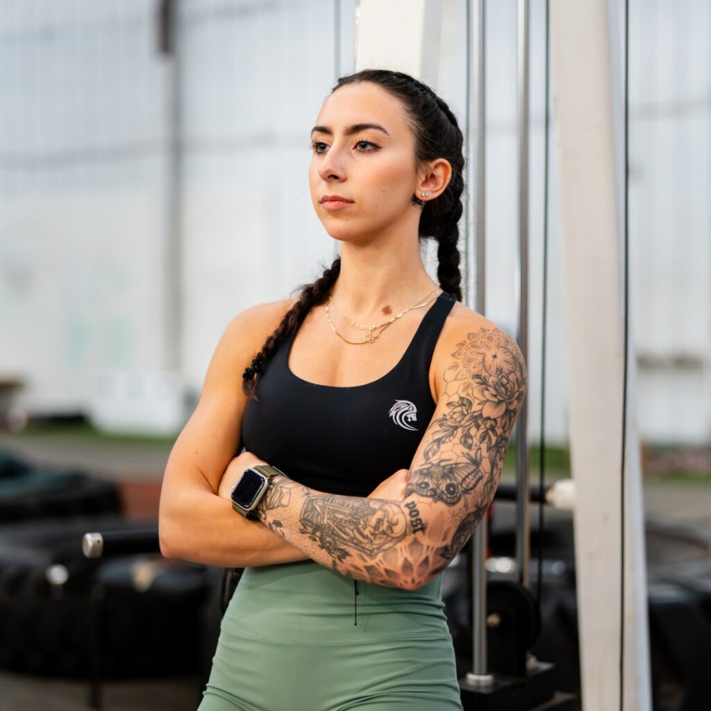 Monthly Brand Content 5 A woman with braided hair and floral tattoos on her arms stands confidently in a gym, wearing a black sports bra and green leggings. She looks focused.