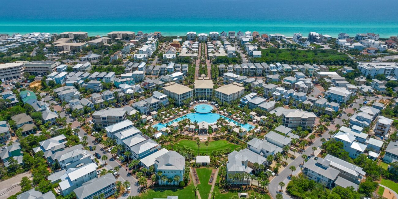 Real Estate 3 Aerial view of a coastal town with a large pool complex at the center, surrounded by neatly arranged houses. The sea sparkles under a blue sky.
