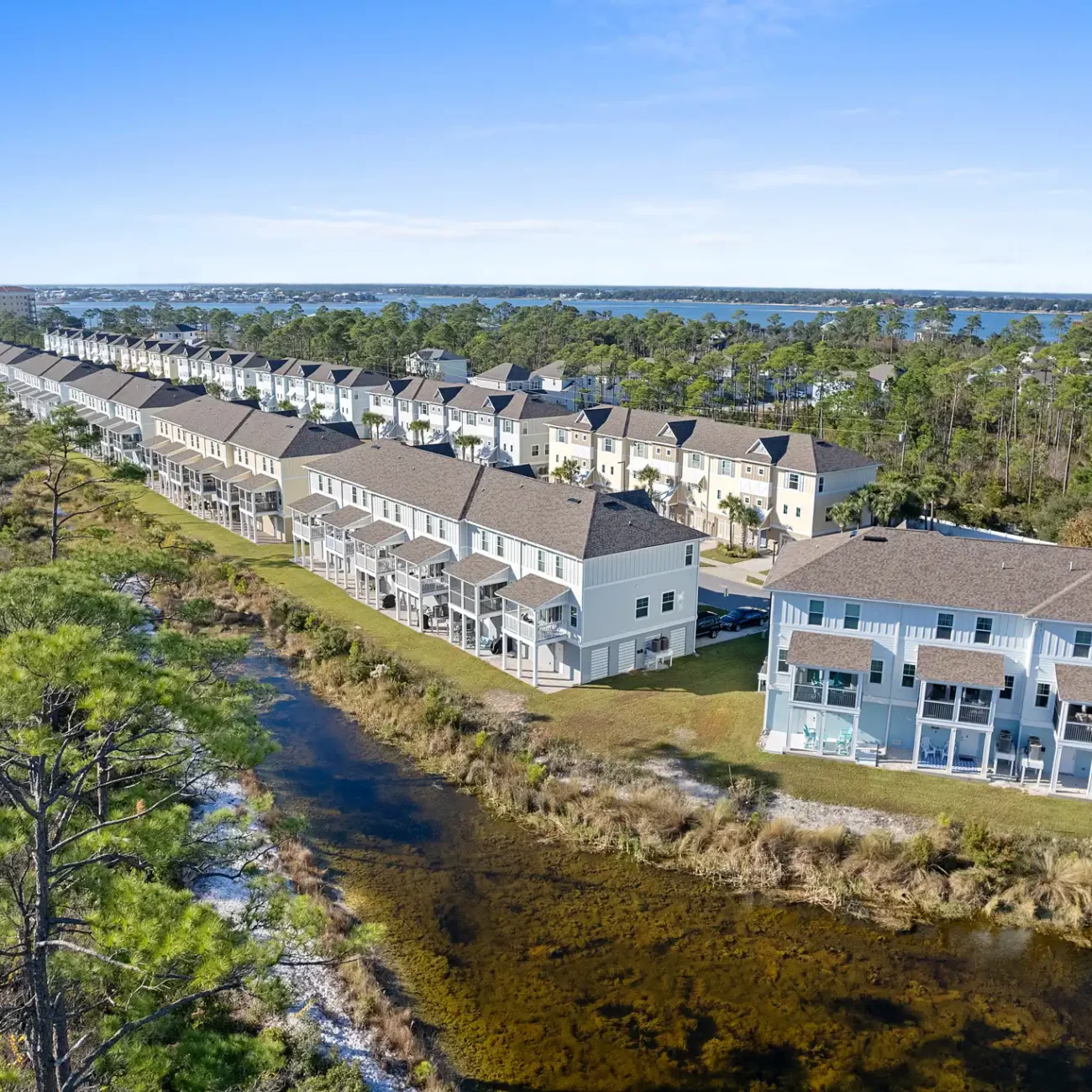 Aerial view of a suburban neighborhood with rows of townhouses bordered by a narrow creek. Lush greenery and a bright blue sky create a serene atmosphere.