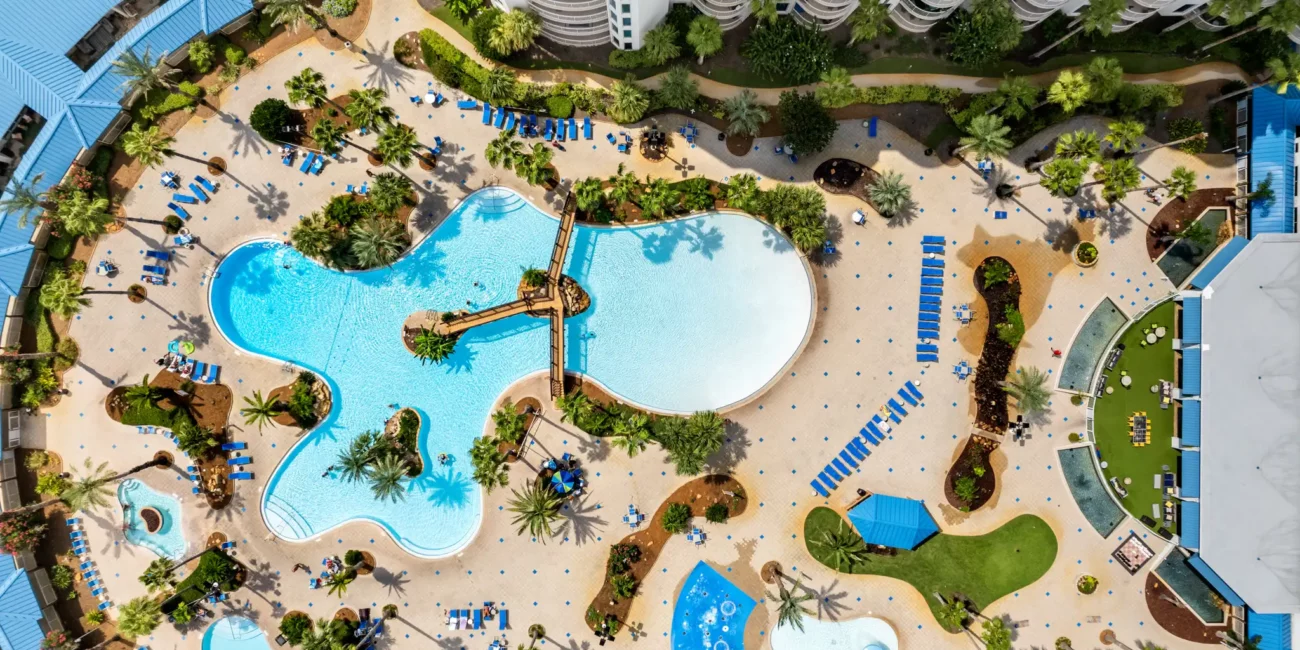 Aerial view of a resort pool area with a large, curvy swimming pool, surrounded by lush greenery and palm trees. Blue loungers line the deck, creating a relaxing atmosphere. Nearby, smaller pools and a bridge add visual interest. The scene is vibrant and inviting.