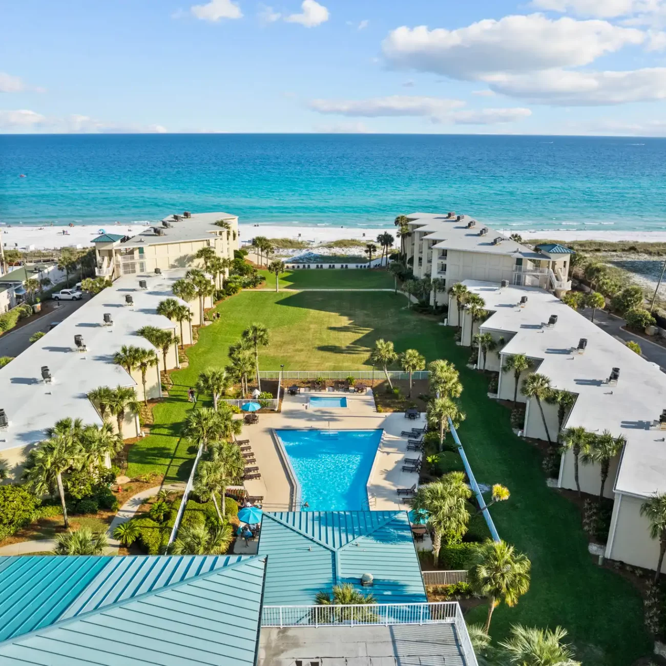 Aerial view of a beachfront resort with a central pool surrounded by palm trees and buildings, overlooking turquoise ocean waves under a blue sky.