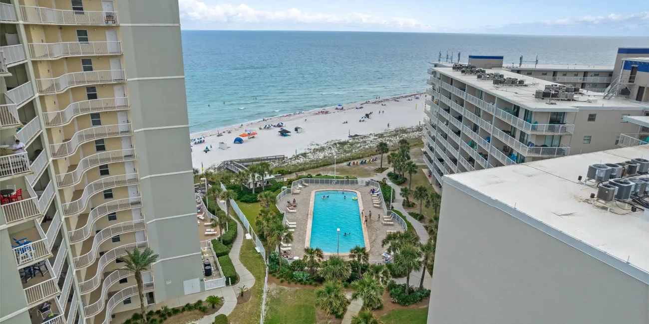 Aerial view of a beachfront condo complex, showing two tall buildings flanking a central pool surrounded by palm trees, with a sandy beach and calm ocean beyond.