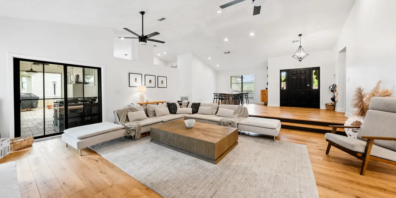 Real Estate 1 Spacious living room with light wooden flooring, large L-shaped beige sofa, wooden coffee table, and modern ceiling fans. Bright and inviting atmosphere.