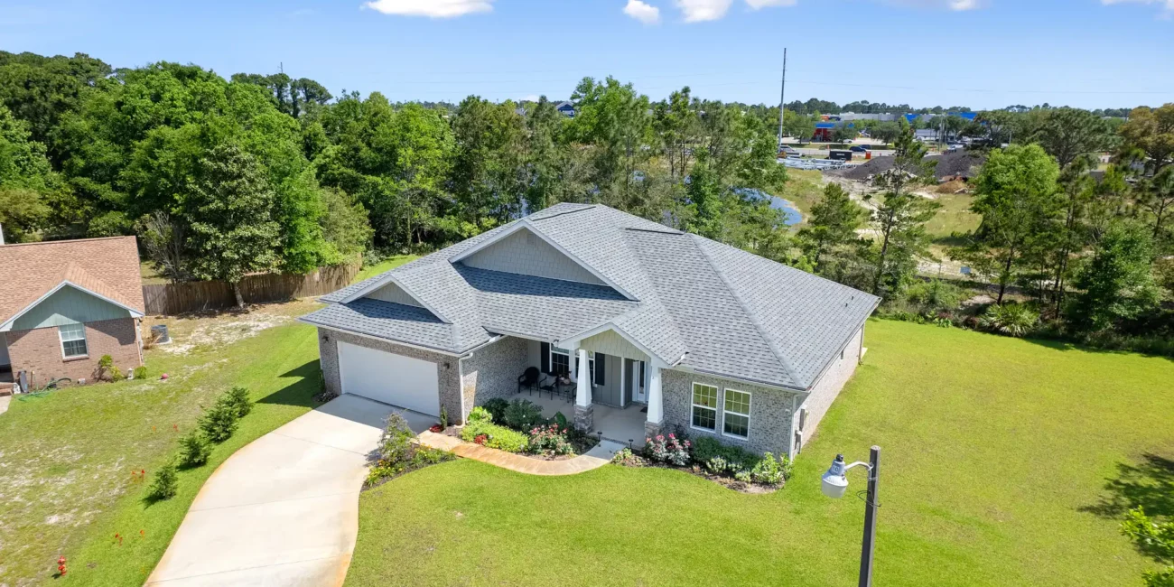 Aerial view of a modern, single-story brick house with a gray roof surrounded by a lush green lawn. The driveway curves to a garage. A few trees and a streetlamp are visible under a blue sky with fluffy clouds.