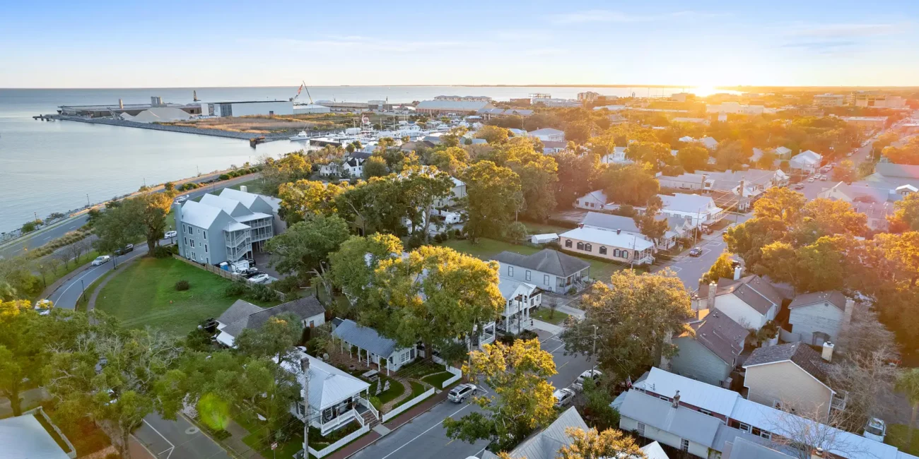 Aerial view of a coastal town at sunset, featuring modern and traditional homes surrounded by lush trees, with a port and ocean in the background.