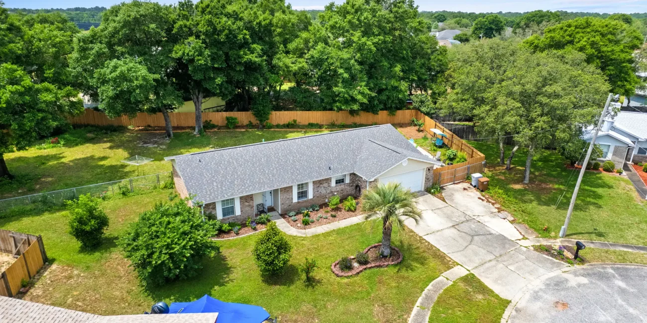 Aerial view of a suburban brick house with a gray roof, surrounded by lush trees and a fenced yard. The driveway leads to a cul-de-sac. Calm, sunny atmosphere.