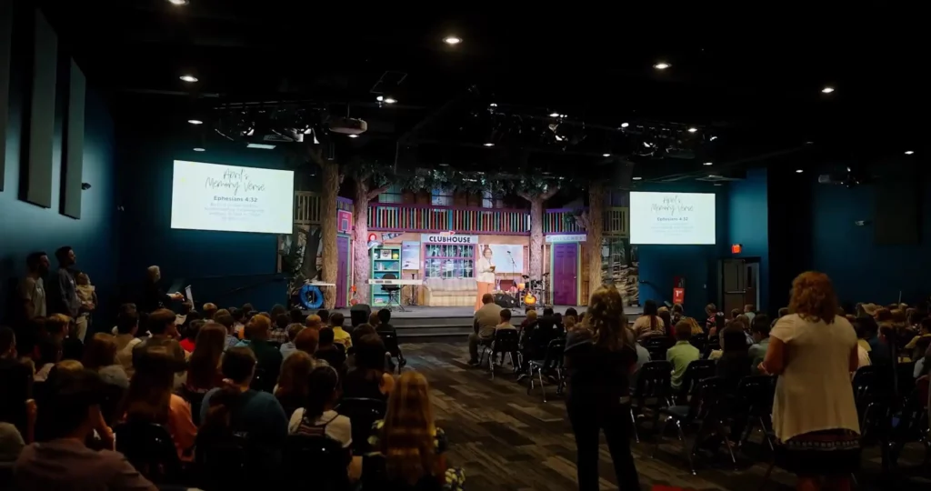 Corporate Event Photography & Videography 3 Wide view of a full auditorium from the back of the room, the audience seated facing a lit stage and two projection screens