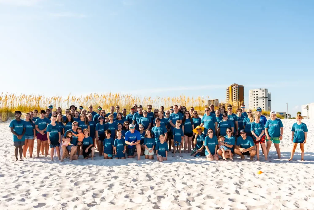Corporate Event Photography & Videography 9 Large group photograph of around a hundred attendees in matching shirts, lined up on the sand with the shoreline behind