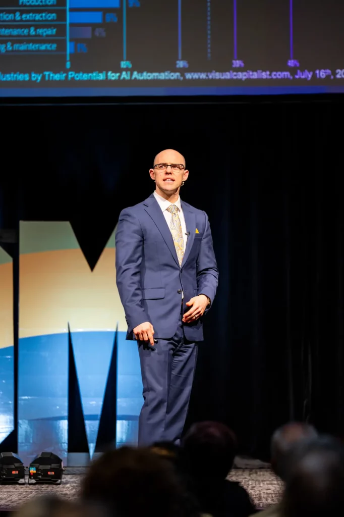 Corporate Event Photography & Videography 1 Keynote speaker in a blue suit alone on a dark stage, a data slide projected above him and the audience in silhouette