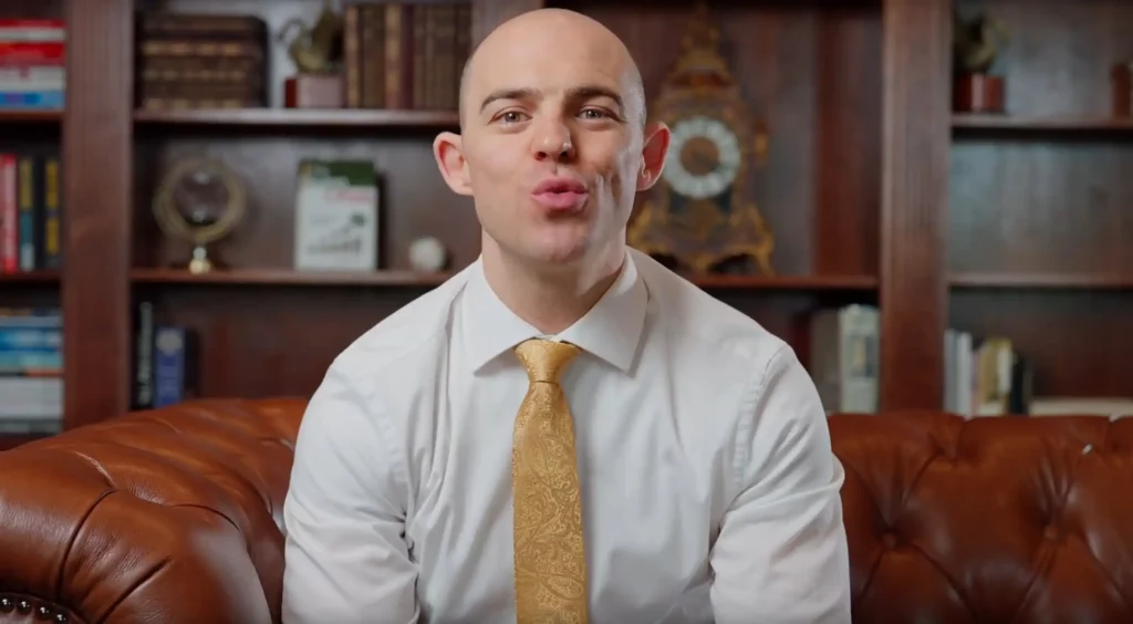 Seated interview set-up in a panelled office, the subject in a white shirt and yellow tie talking to camera