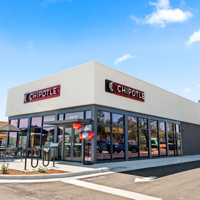 Chipotle Mexican Grill at the Ransley Station development 8870 Boesch Ln Pensacola FL A Chipotle restaurant with glass windows and doors, adorned with red and silver balloons. Outdoor seating is visible under a clear blue sky.