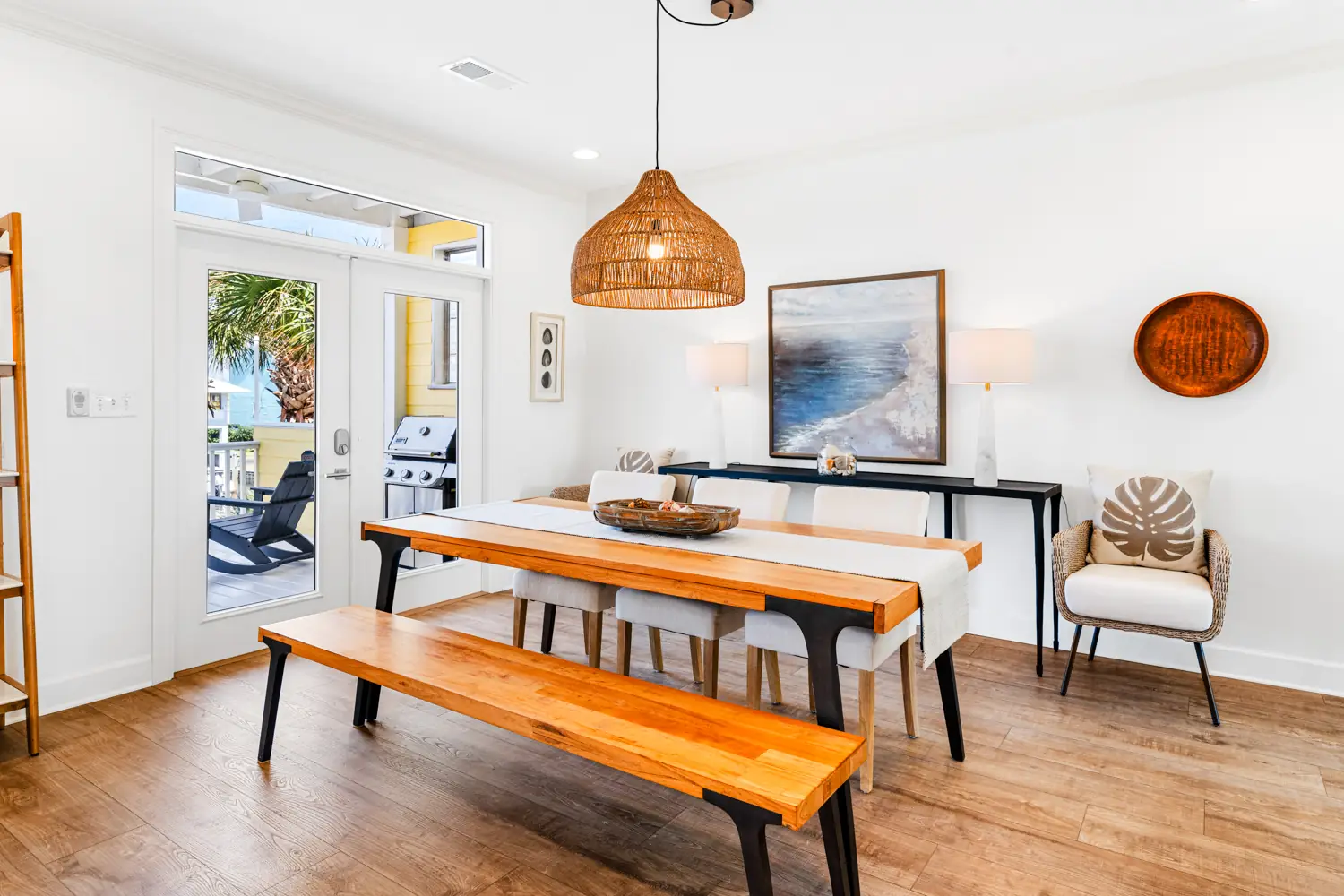 Real Estate Photography 1 Bright dining room with a wooden table and bench, wicker light fixture, coastal artwork, and a view of a patio with a grill in the background.