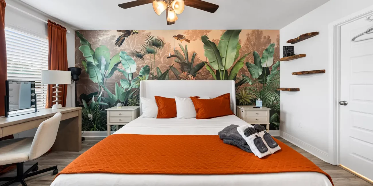 Cozy bedroom with tropical wall mural, featuring a white bed with orange accents, wooden shelves, a desk, and a ceiling fan, creating a warm, inviting ambiance.