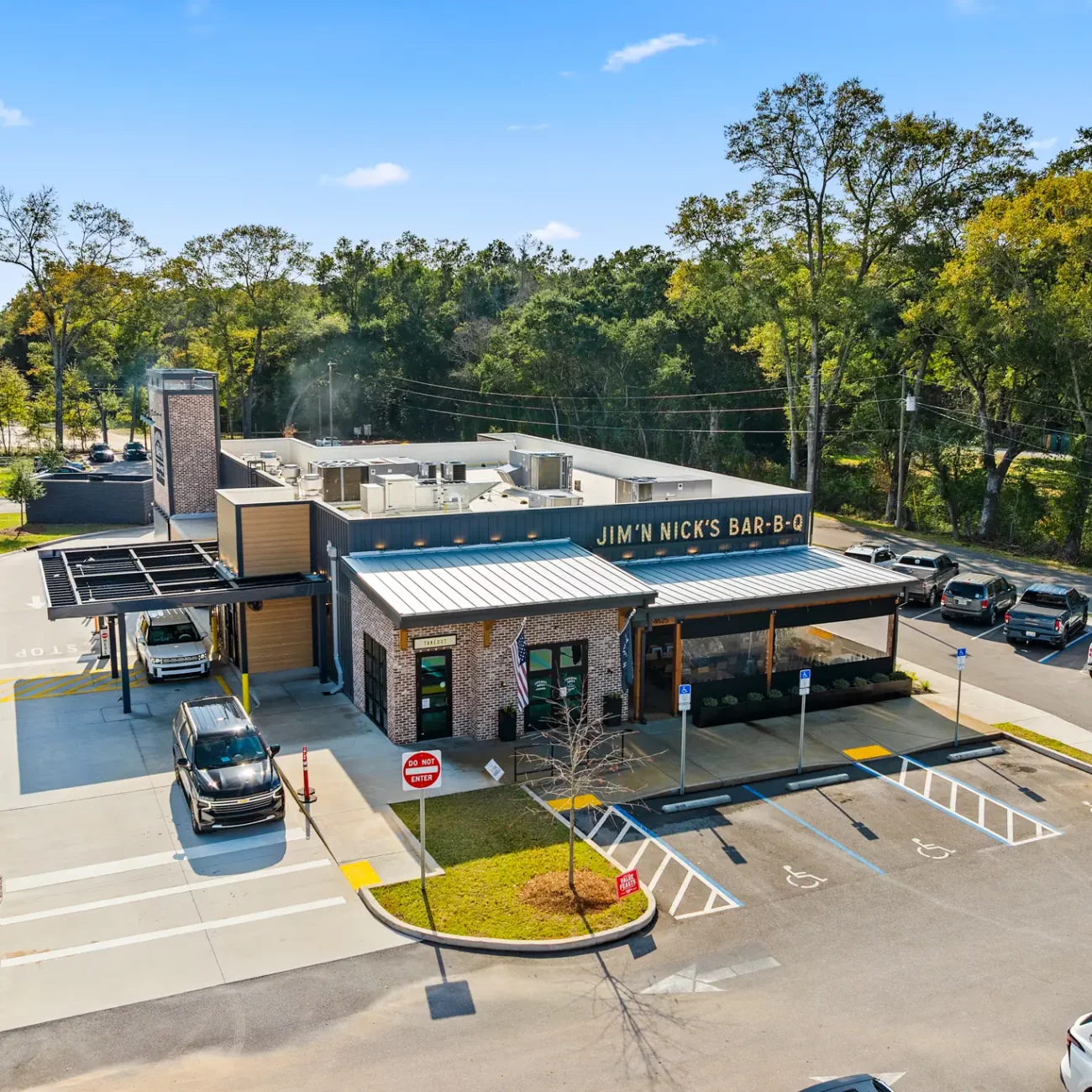 Jim-N-Nicks Pace FL Aerial view of a barbecue restaurant with a modern design, surrounded by parked cars. Greenery and trees in the background under a clear blue sky.